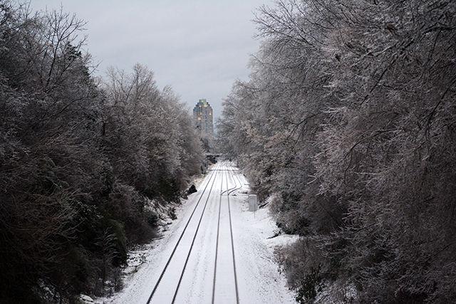Overlooking Pullen Road, the rail track is covered in snow and there is a glimpse of downtown in between all the white trees.