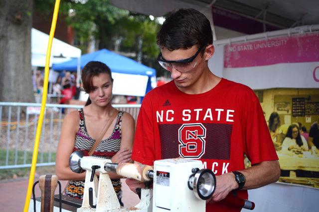 Colin Moore, a freshman studying engineering, constructs a wooden bud vase with a lathe machine while his girlfriend Emily Tatum looks on at the Crafts Center booth in the Arts Zone on Hillsborough Street during Packapalooza on Aug. 20. Representatives from the Crafts Center helped participants use a wood lathe machine to make their own wooden flower vases.