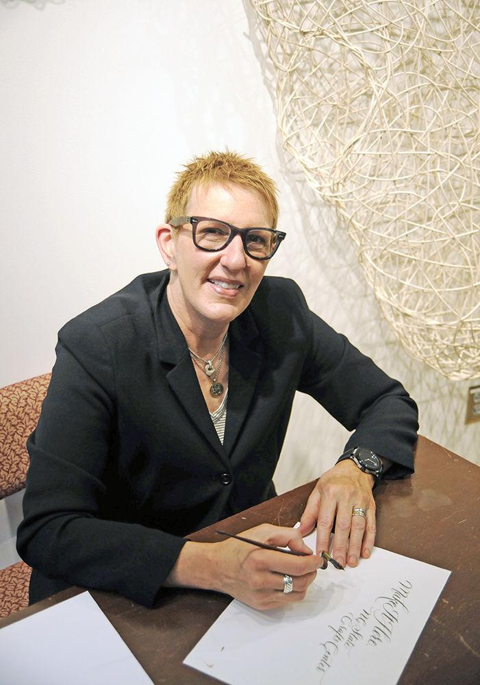Director of the NC State Crafts Center Carol Fountain Nix poses for a portrait with her calligraphy pen in front of a free weaving sculptural installation by artist Anne Wilson, which serves as a centerpiece for the craft center. The words written on her page are: "Make it Here, The NC State Crafts Center,&#8221; a widely used slogan of the the center.