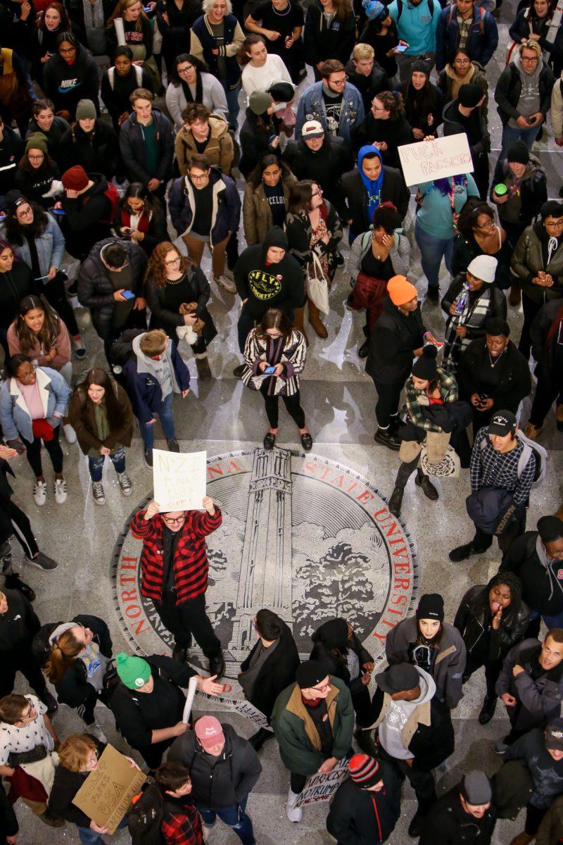Garrett Justice, a senior studying Sociology, holds a protest sign while standing on the NC State seal during the No Hate At NC State protest of a Turning Point event in Talley Student Center on Wednesday, Nov. 13, 2019. "I've got a lot of friends who've been affected by the policies of the Trump administration," he said. "I'm here to fight that, and hopefully make a change."
