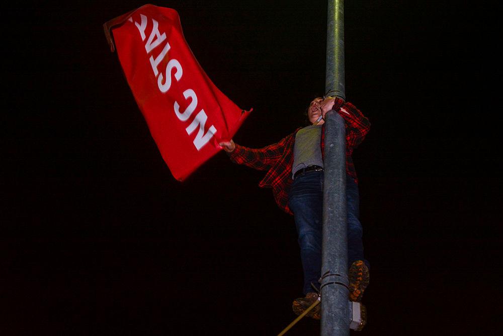 An NC State student climbs a light pole and waves an NC State &#8220;Think and Do&#8221; banner above the crowd. NC State students rushed the Bell Tower after the 88-66 win over Duke on Wednesday, Feb. 19, 2020. Hillsborough Street flooded with Wolfpack fans in support of the men&#8217;s basketball team win.