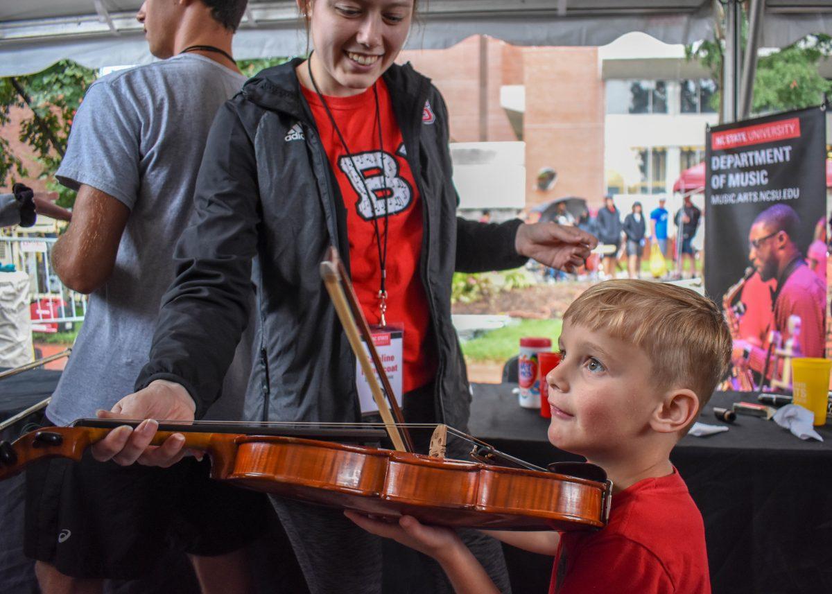 Emerson Kasopsky holds a violin while Department of Music member Caroline Heathcoat teaches him how to play during Packapalooza on Saturday, Aug. 24, 2019 on Hillsborough Street. The 2019 Packapalooza event is a continuation of NC State's annual block-party tradition to ring in the new school year with vendors, artists, live performances, food and giveaways.