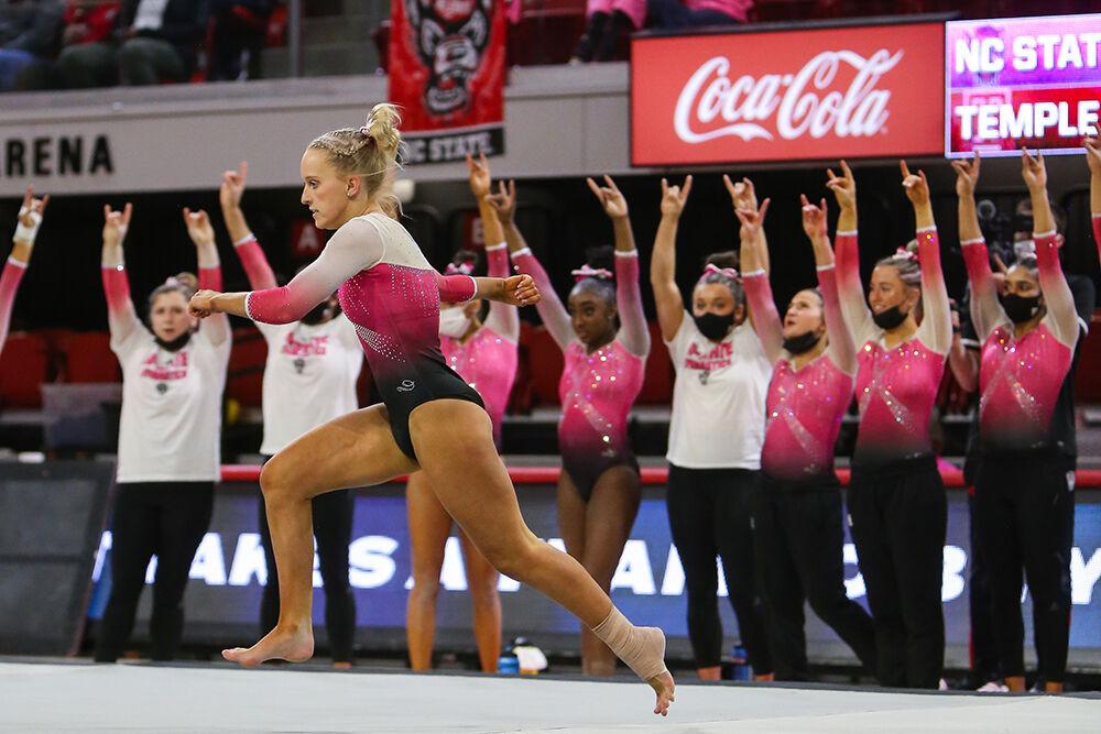 The NC State gymnastics team holds up their Wolfpack "wolfies" while freshman Brooke Smead goes to perform a front handspring on the floor at the Pink Meet against Temple on Friday, Feb. 25, 2022. NC State lost 196.675-196.600.