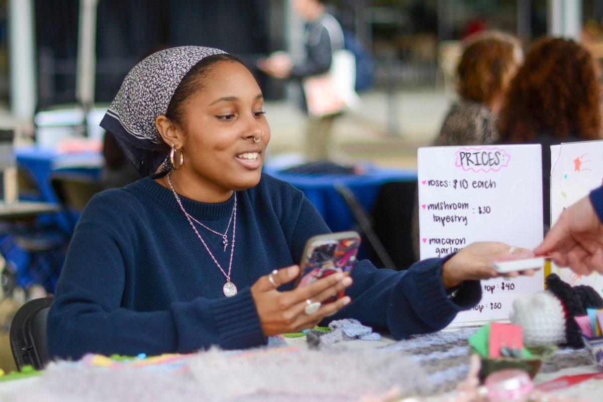 Havilynn Mills, a third-year in design studies, smiles as she checks out a customer at the CYC Small Business Fair on Stafford Commons on Tuesday, April 9, 2024. Mills' business, Handmade by Havilynn, offers crochet tapestries, roses and more.